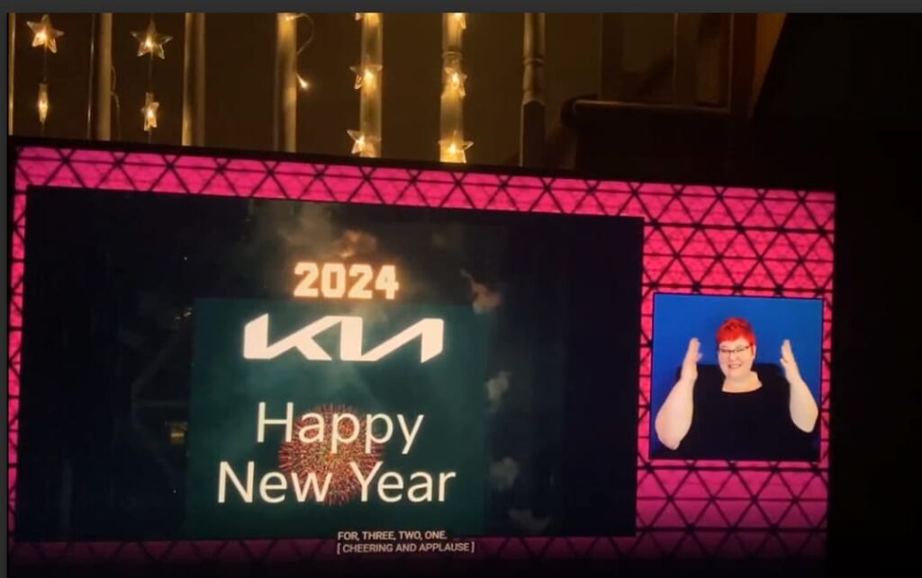 The YouTube livestream on a screen at home: a 'Happy New Year 2024' graphic in the main frame, the ASL interpreter in a panel on the right, and the caption 'For, three, two, one. [Cheering and applause]' below.