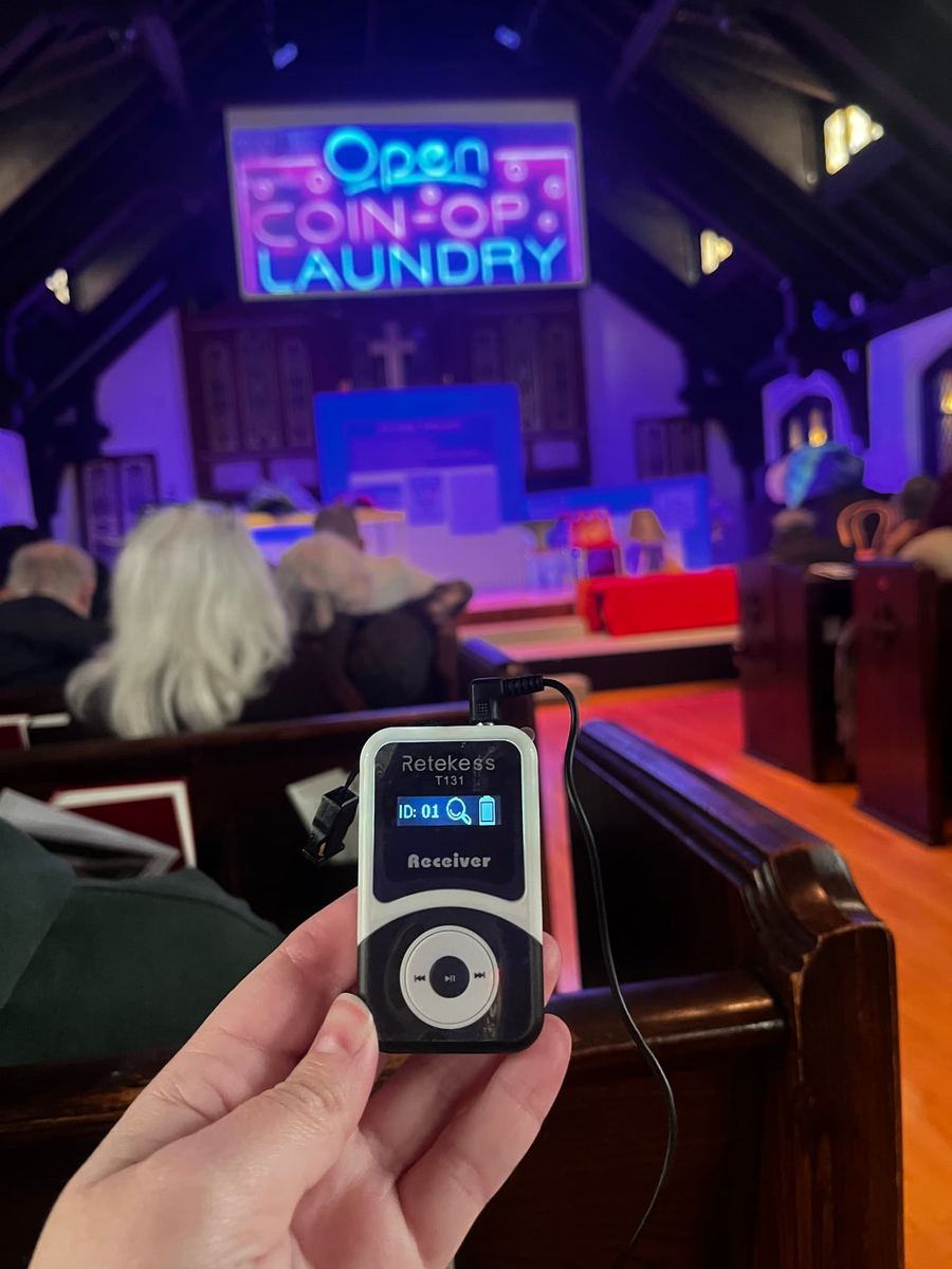 The theatre stage is in a modern church with colorful lights. There is a neon "Open Coin-Op Laundry" sign and an attendee with multilingual headset audio receiver.