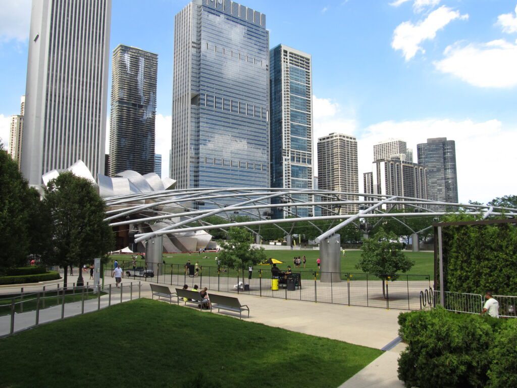 The curving steel trellis stretched over the lawn at Millennium Park, the stainless-steel bandshell of the Pritzker Pavilion at the left edge and downtown towers rising behind, with a few people sitting on benches outside the fence.