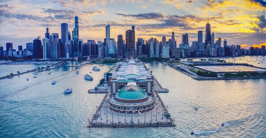 Navy Pier photographed from the air at sunset, the green-domed ballroom at the far end of the pier, the Ferris wheel and exhibition halls behind it, and the downtown skyline running along the lakeshore.