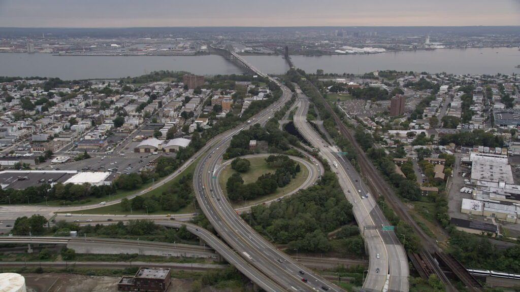 An aerial view of a cloverleaf highway interchange and a rail line cutting through a dense low-rise neighborhood, with a long bridge crossing open water toward a distant skyline.