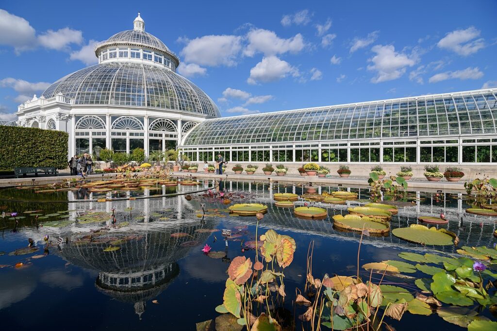 The domed glass Palm House of the New York Botanical Garden's conservatory, reflected in the lily pond in front of it, with giant lily pads floating in the foreground.