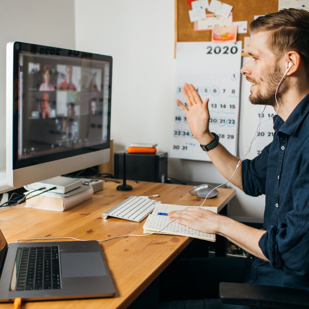 A man waves at a video call grid of colleagues on his home computer.