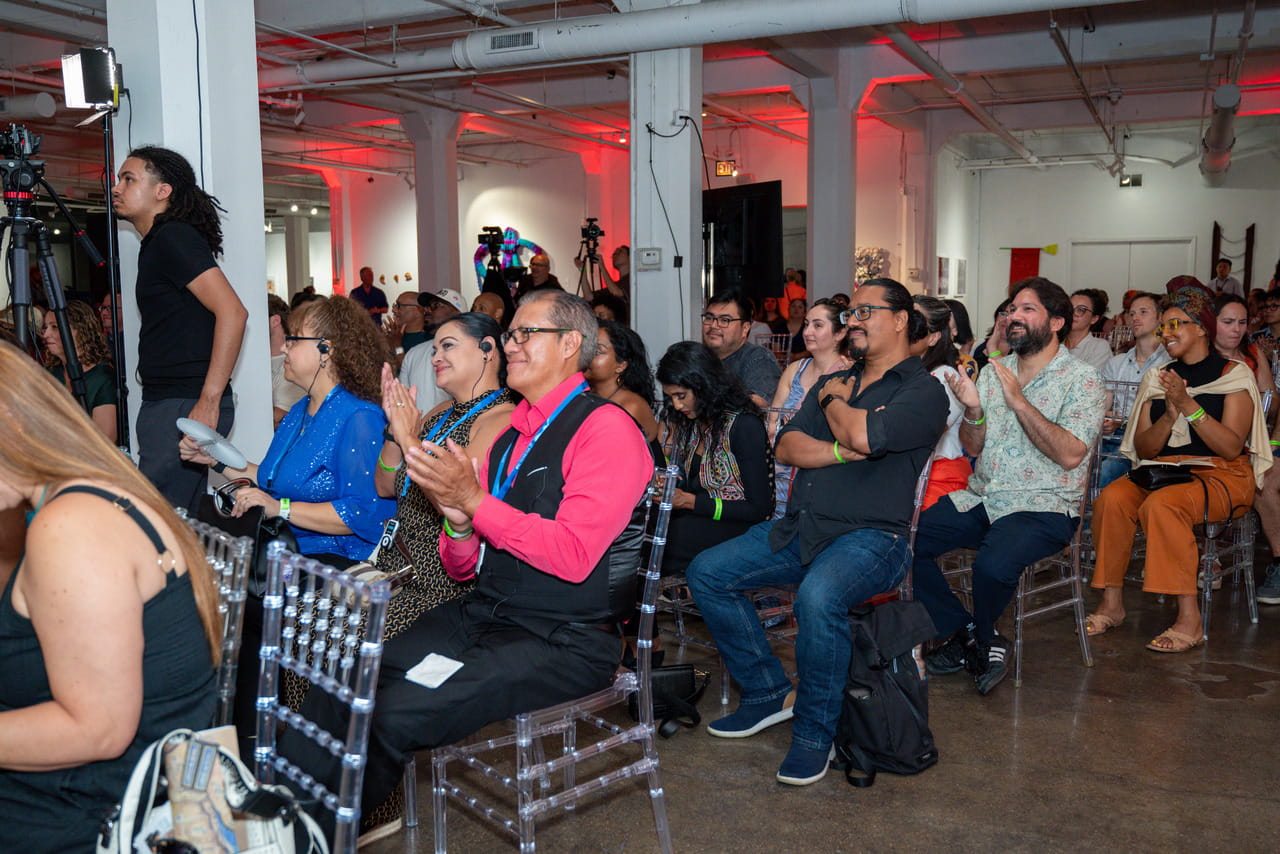 The audience at the Chicago event applauding from clear acrylic chairs in a gallery space, several attendees in the front rows wearing lanyards and single earpieces carrying the Spanish interpretation.