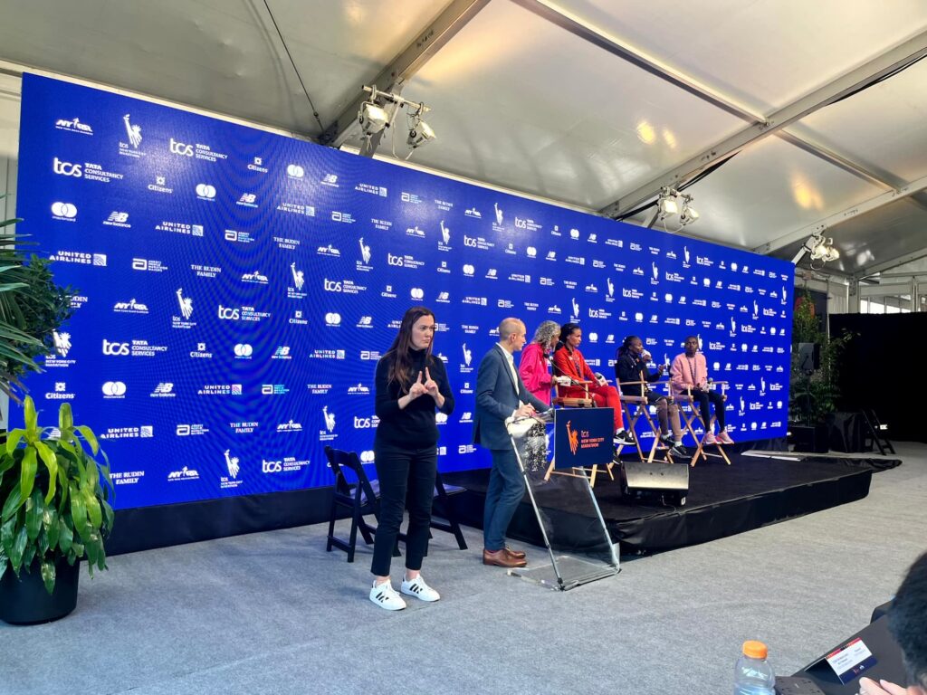 An ASL interpreter signs at the front of a marathon press conference while elite runners sit for a panel on stage in front of a blue TCS New York City Marathon sponsor backdrop.