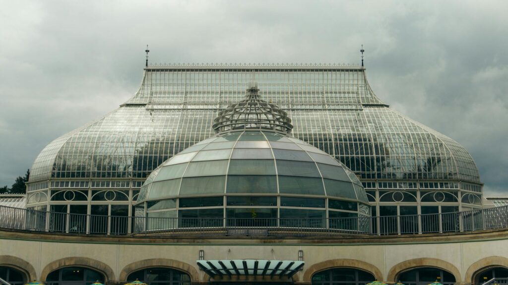 The glass barrel roof and central dome of the Phipps Conservatory palm house under a heavy grey sky, its railed terrace sitting above an arcaded stone base.