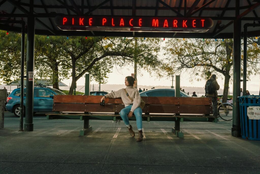 A woman sits on a long wooden bench under a shelter, a red neon sign reading Pike Place Market above her, a man with a backpack standing at the right, trees still in yellowing autumn leaf and parked cars behind, and water in the distance.