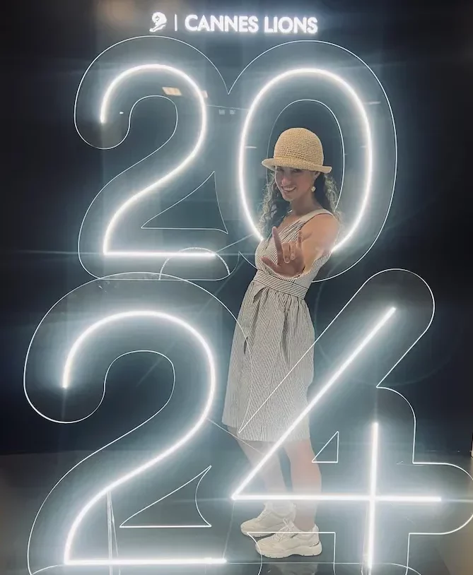 Rebecca stands in front of a Cannes Lions neon-lit '2024' display, holding up the ILY hand sign.