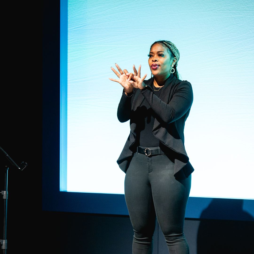 A woman signs on stage in front of a large projection screen.