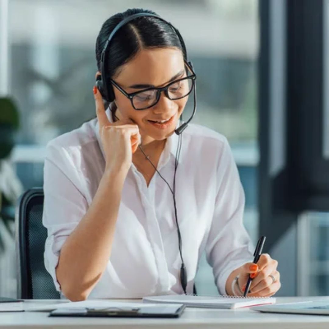 A woman wearing a headset takes notes at her desk.