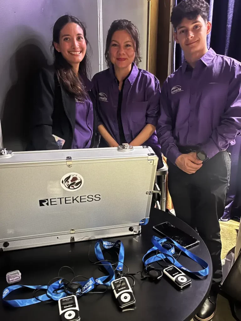 Three ICS team members in purple Borealis shirts stand behind an open Retekess equipment case, with interpreting receivers and headsets laid out on the table in front of them.