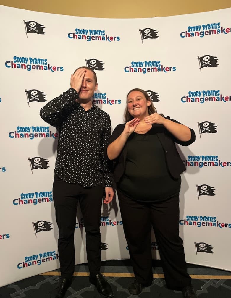 The co-founders of ICS, Shelby and Tyler, are standing behind a banner that reads “Story Pirates Changemakers.” The banner background is white, and the words are printed in multiple colors. Shelby is signing ‘story’ and Tyler is signing ‘pirate.’