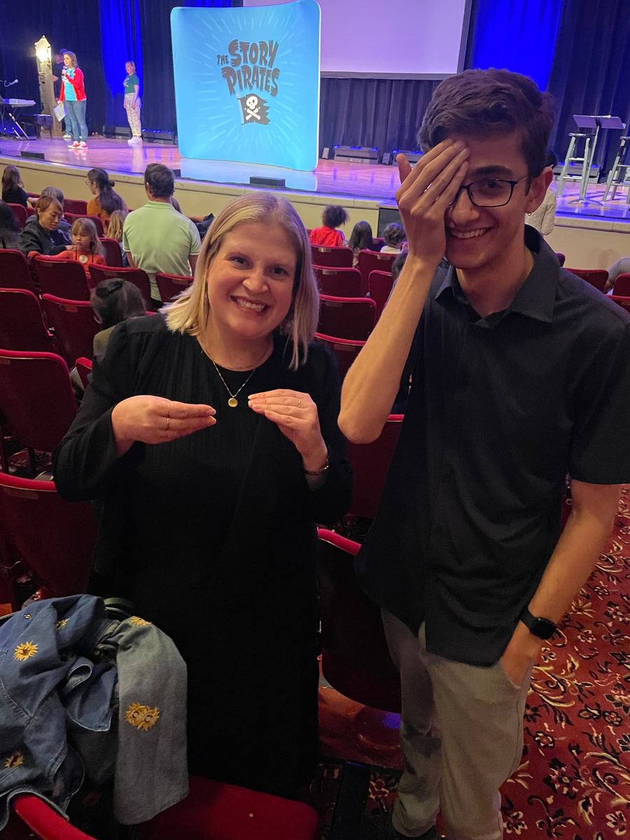 Two people standing in the back of the theatre. A woman is signing ‘story’ while a young man wearing glasses is signing ‘pirate.’