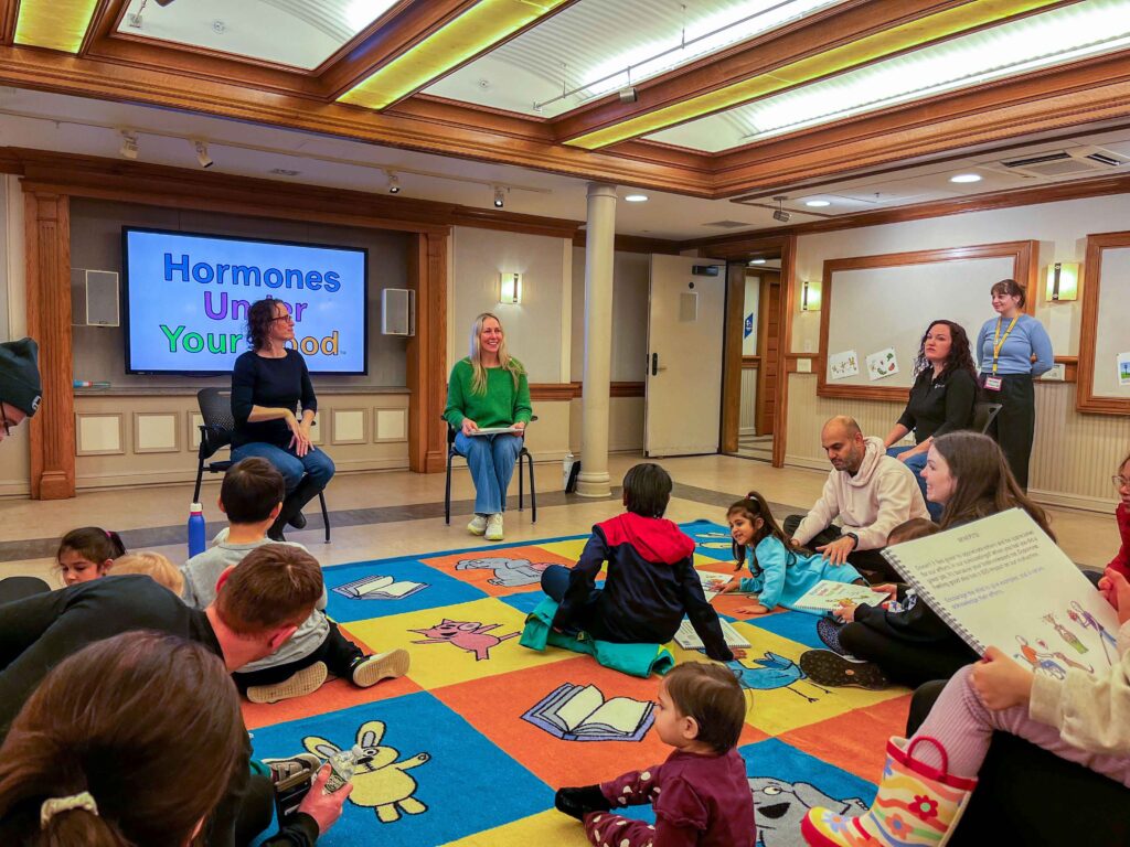 A story hour in a library reading room: children and parents sit on a patterned rug while the author and the Deaf interpreter read from chairs at the front, the Hormones Under Your Hood title on the screen behind them.