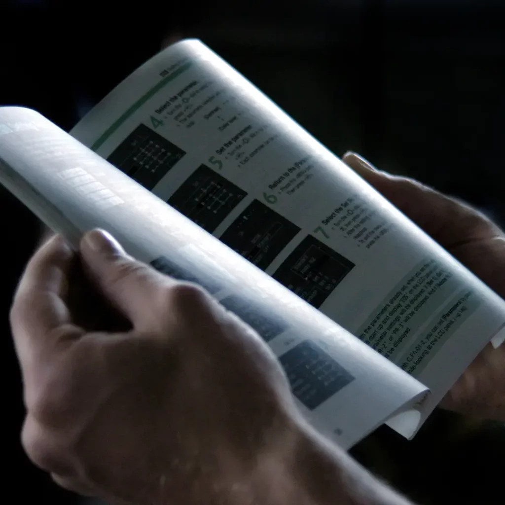 The illustration the source page used for this section: two hands holding open a printed technical manual of numbered step-by-step instructions, lit against a dark background.