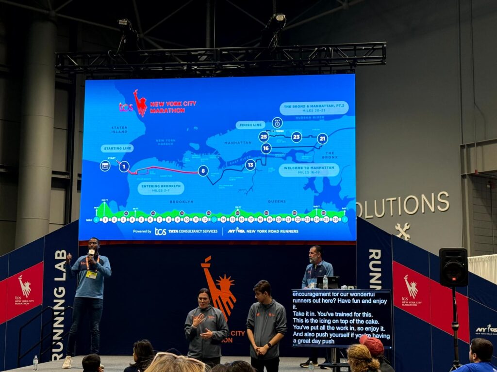A course strategy session at the expo. A large screen shows the TCS New York City Marathon route map through the five boroughs while speakers present below it; the ASL interpreter team is pictured at the bottom right of the photo, beside a monitor displaying live captions.