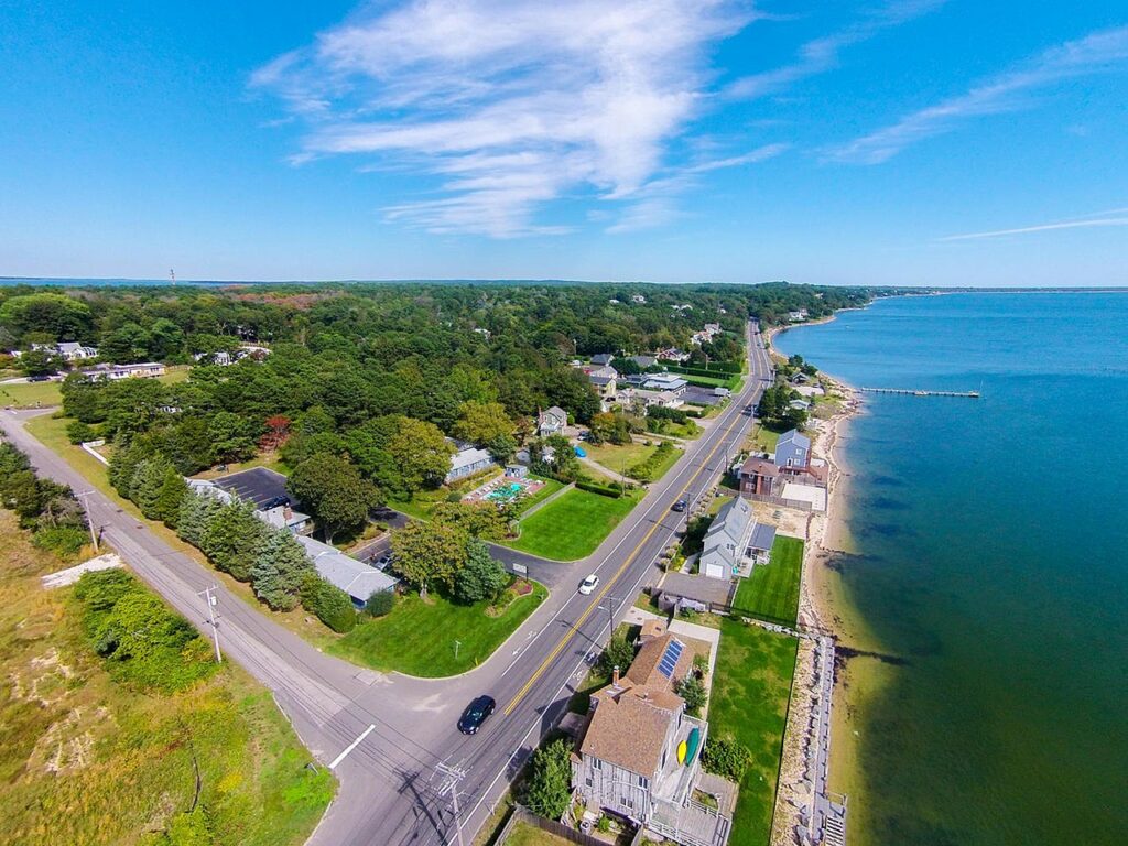 An aerial view of a tree-lined shore road running past houses and lawns along the bay, with a narrow pier reaching into the water, in one of the Hamptons villages on Long Island's East End.