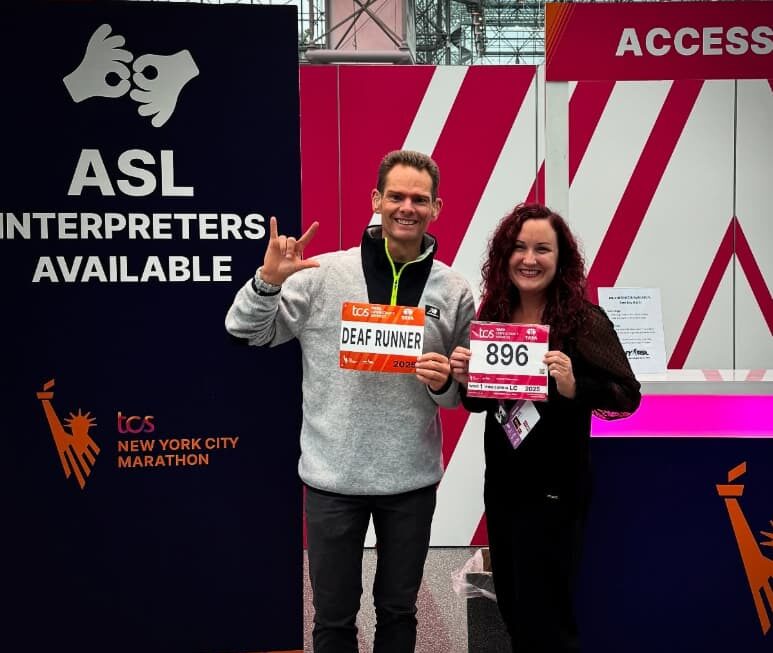 Thomas Eller holds up a 'Deaf Runner' race bib and the ILY handshape beside Sarah, who holds bib number 896, in front of an 'ASL Interpreters Available' sign at the expo accessibility area.