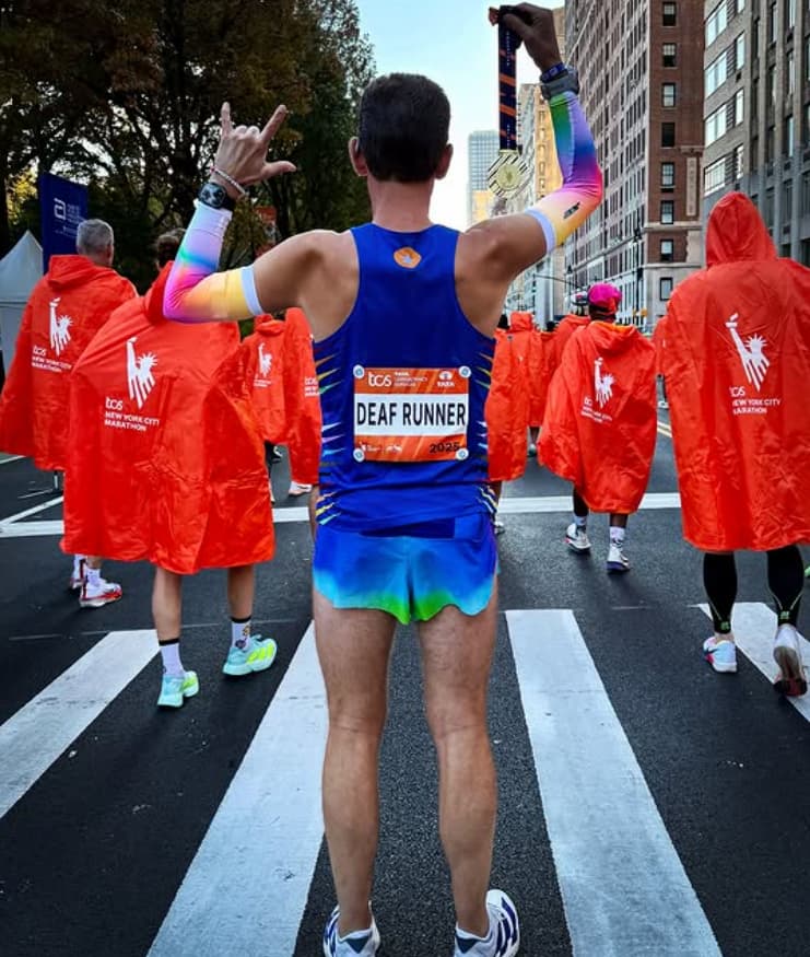 Thomas Eller, seen from behind in a blue singlet with a 'Deaf Runner' race bib, raises his medal in one hand and the ILY handshape in the other while walking through the post-race area among runners in orange ponchos.