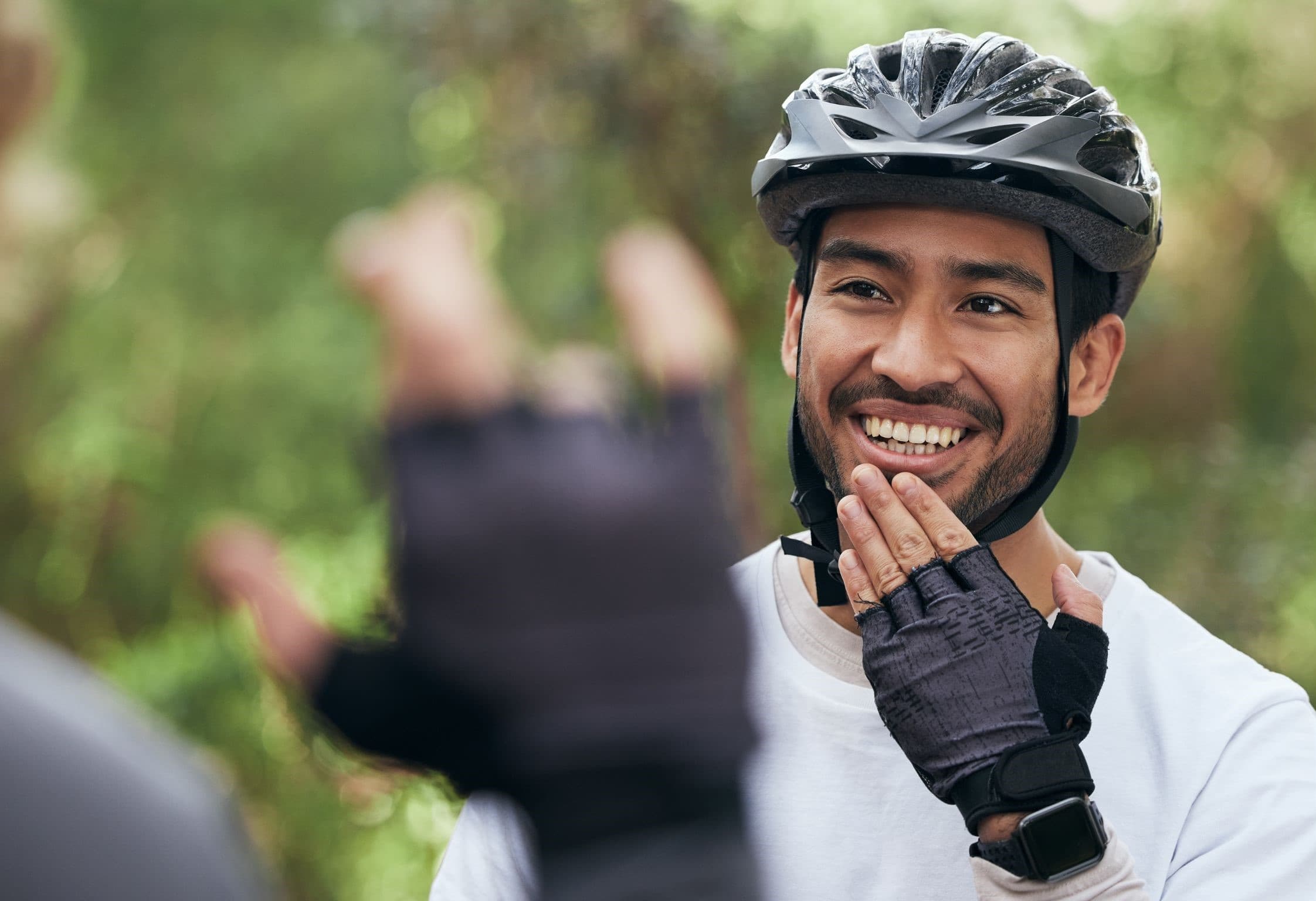 A smiling cyclist in a helmet watches someone sign outdoors on a wooded trail