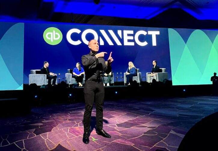 An ASL interpreter signs centre stage in front of the QuickBooks Connect backdrop while five panelists sit in armchairs behind him.