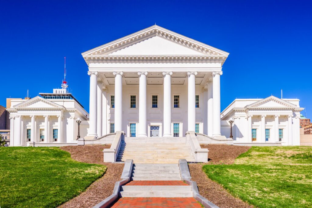 The Virginia State Capitol's white Roman-temple front, its portico of six columns above a flight of steps cut into the lawn, with a lower pedimented wing standing on either side.
